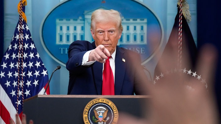 Das Bild zeigt Donald Trump während einer Pressekonferenz wie er auch eine Person zeigt.