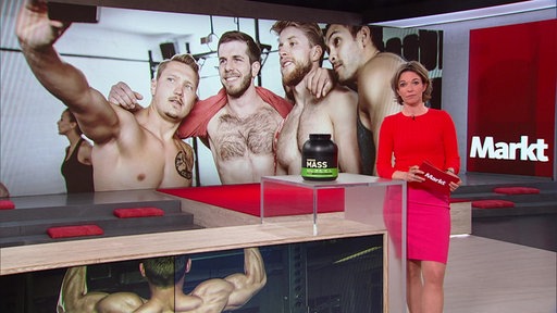 Anna Planken steht im Markt-Studio. Auf dem Display hinter ihr, wird eine Gruppe von jungen Männern angezeigt.