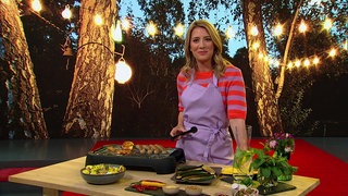 Catherine Vogel steht im Markt-Studio.  Auf dem Moderationstisch sieht man ein gestelltes Grill-Set, mit viel Gemüse und Fleisch. Im Hintergrund sieht man eine Abbildung eines Gartens mit Lichterkette.