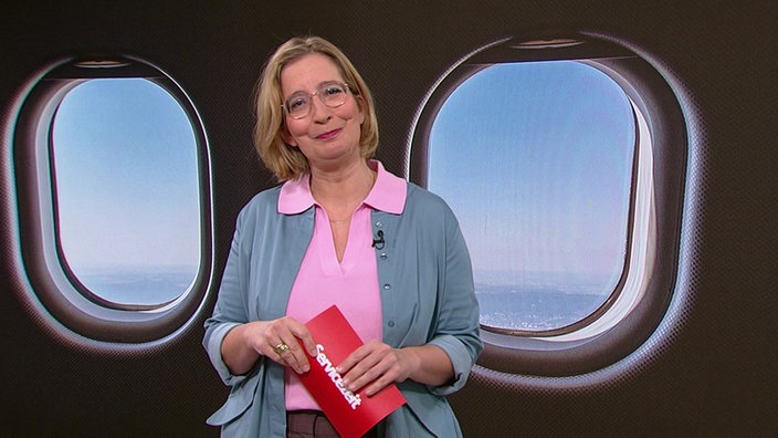 Yvonne Willicks steht im Servicezeit-Studio.  Im Hintergrund sieht man den Innenraum eines Flugzeuges.