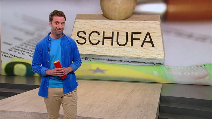 Daniel Aßmann steht im Servicezeit-Studio. Im Hintergrund sieht man das Bild eines Stempels mit der Betitelung "SCHUFA".