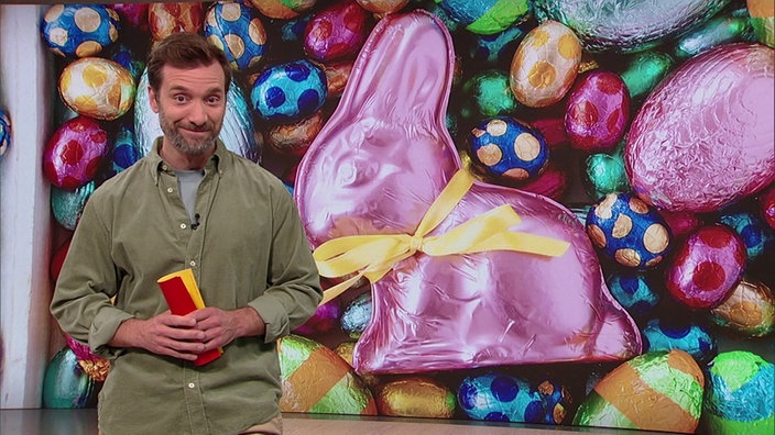 Daniel Aßmann steht im Servicezeit-Studio. Im HIntergrund siehz man verschiedenen Osterschokoladen-Produkte präsentiert.