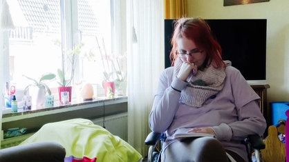 Annika Witteler sitzt in ihrem Wohnzimmer vor einem Fernseher, neben einem großen Fenster. Sie stützt ihren Kopf nachdenklich auf ihrem rechten Arm ab. 