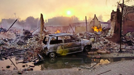 Ein ausgebranntes Auto steht vor Gebäudetrümmern. Im Hintergrund lodern weiter Flammen.