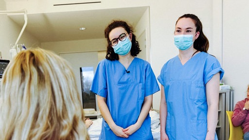 Lara Soyubey und Xenia Look stehen gemeinsam vor einer Patientin. Die beiden jungen Frauen tragen beide einen Mundschutz.