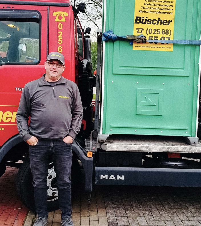 Berni Bösing, ein Mann mit Brille und Kappe steht vor einem roten Reinigungsfahrzeug für Toiletten. Auf dem Fahrzeug ist eine grüne Toilettenkabine mit der Aufschrift "Büscher".