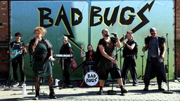 Die Inszenierung Bad Bugs des Theater Marabu in Bonn. Auf der Bühne stehen Menschen in kostümen, Sie singen in Mikrofone und spielen Instrumente. Im Hintergrund steht Bad Bugs auf einer Wand. | Bildquelle: Theater Marabu/Ursula Kaufmann Die Inszenierung Bad Bugs des Theater Marabu in Bonn. Auf der Bühne stehen Menschen in kostümen, Sie singen in Mikrofone und spielen Instrumente. Im Hintergrund steht Bad Bugs auf einer Wand.