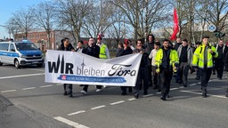 Ein Demozug mit teils in Warnwesten bekleideten Ford-Mitarbeitern zieht an einem Polizeiwagen vorbei. Vorne haben sie ein Banner mit der Aufschrift "Wir bleiben Ford" | Bildquelle: WDR / Piotrowski Ein Demozug mit teils in Warnwesten bekleideten Ford-Mitarbeitern zieht an einem Polizeiwagen vorbei. Vorne haben sie ein Banner mit der Aufschrift "Wir bleiben Ford"