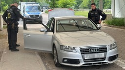 Auf dem Bild ist ein grauer Wagen und mehrere Polizisten. Im Hintergrund ist ein Polizeibulli.