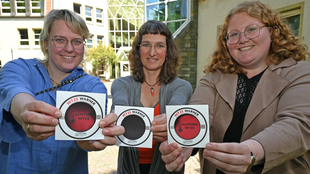 Drei Frauen die Sticker mit der Aufschrift "Hitze Warner" ins Bild halten.