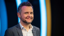 Moderator Tobias Mann steht während der Verleihung des 50. Deutschen Kleinkunstpreises 2022 im Frankfurter Hof auf der Bühne und lächelt in die Kamera. 