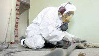Das Bild zeigt eine Person in einem Schutzanzug und einer Gasmaske, die ein Gebäude saniert.